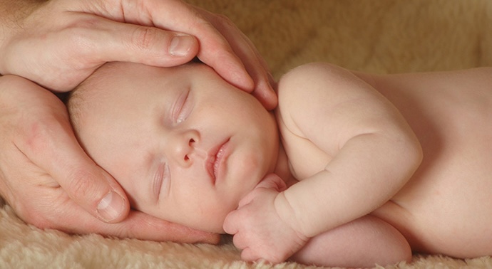 A baby with eyes closed being treated with two hands holding the head